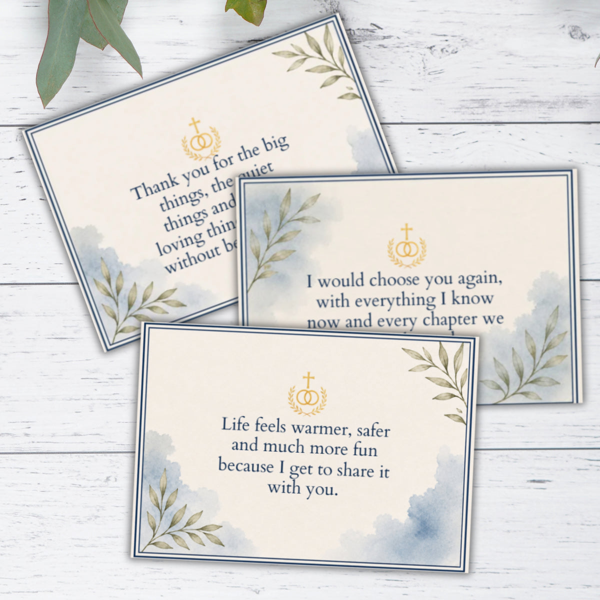 Three wedding invitation cards with elegant watercolor blue-gray leaf designs and gold monogrammed crosses, arranged on a white wood background.