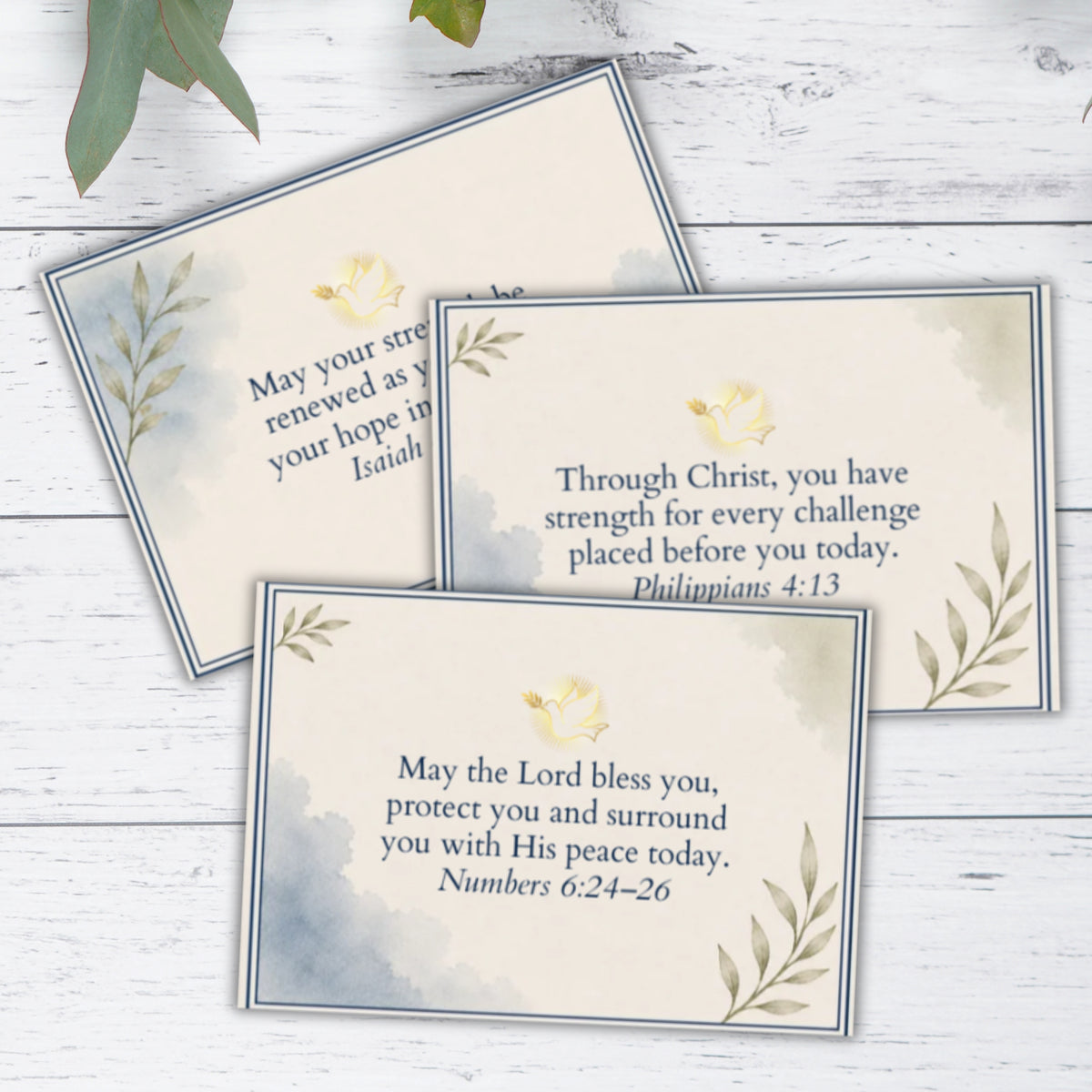 Three Bible verse prayer cards with watercolor blue-gray backgrounds, navy borders, and small yellow floral illustrations, arranged on a white wood surface with green leaves at the top.