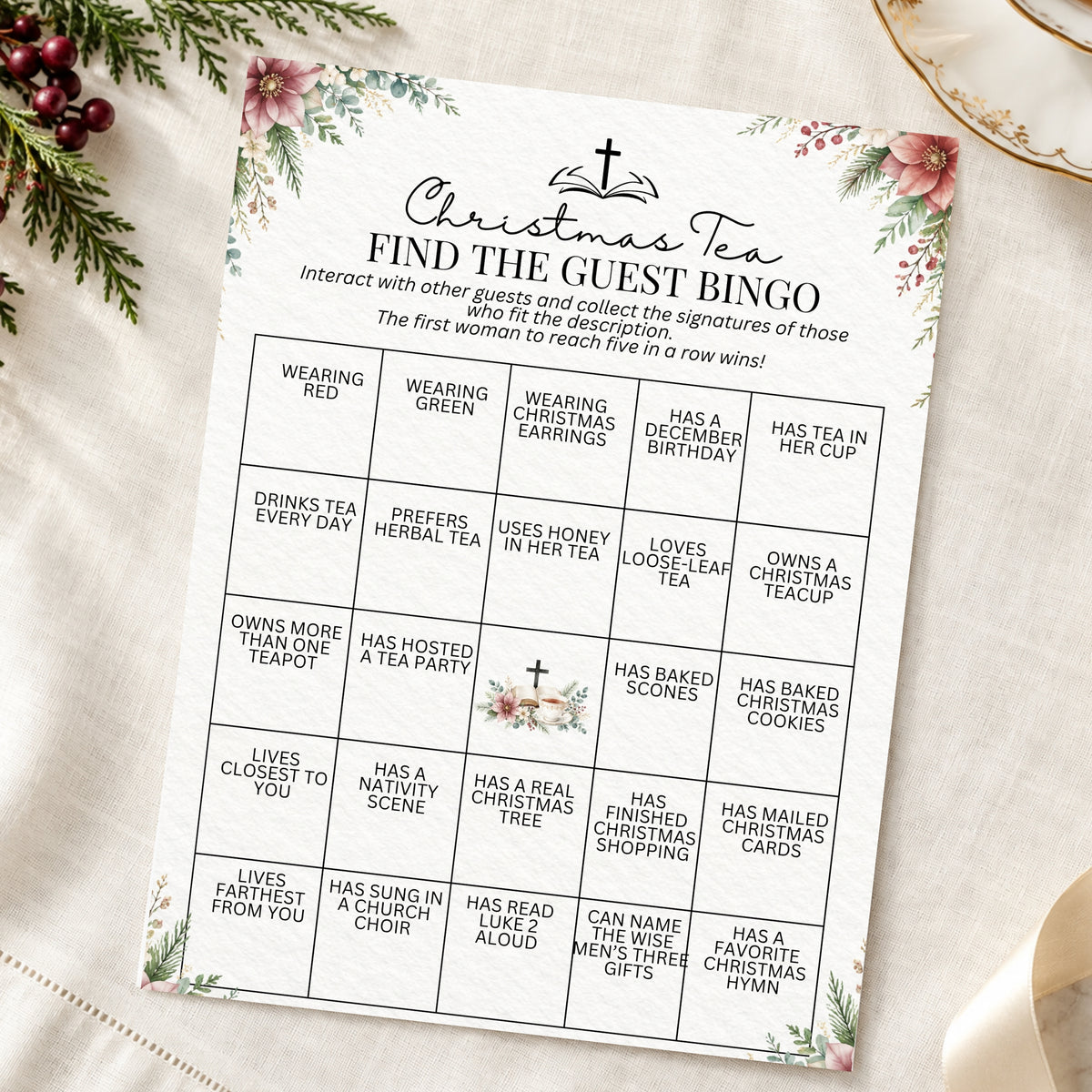 A Christmas-themed 'Find the Guest Bingo' card with floral borders, photographed flat on a cream fabric surface with holiday greenery and a gold plate in the background.