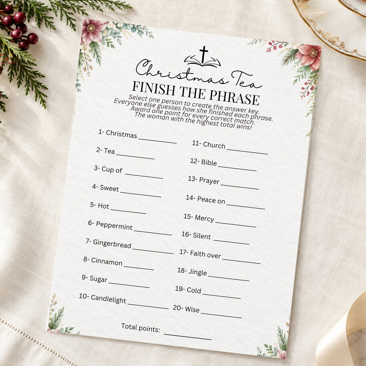 A Christmas-themed 'Finish the Phrase' game card printed on white paper with floral corner designs, photographed on a cream fabric surface with holiday greenery and a gold plate as props.
