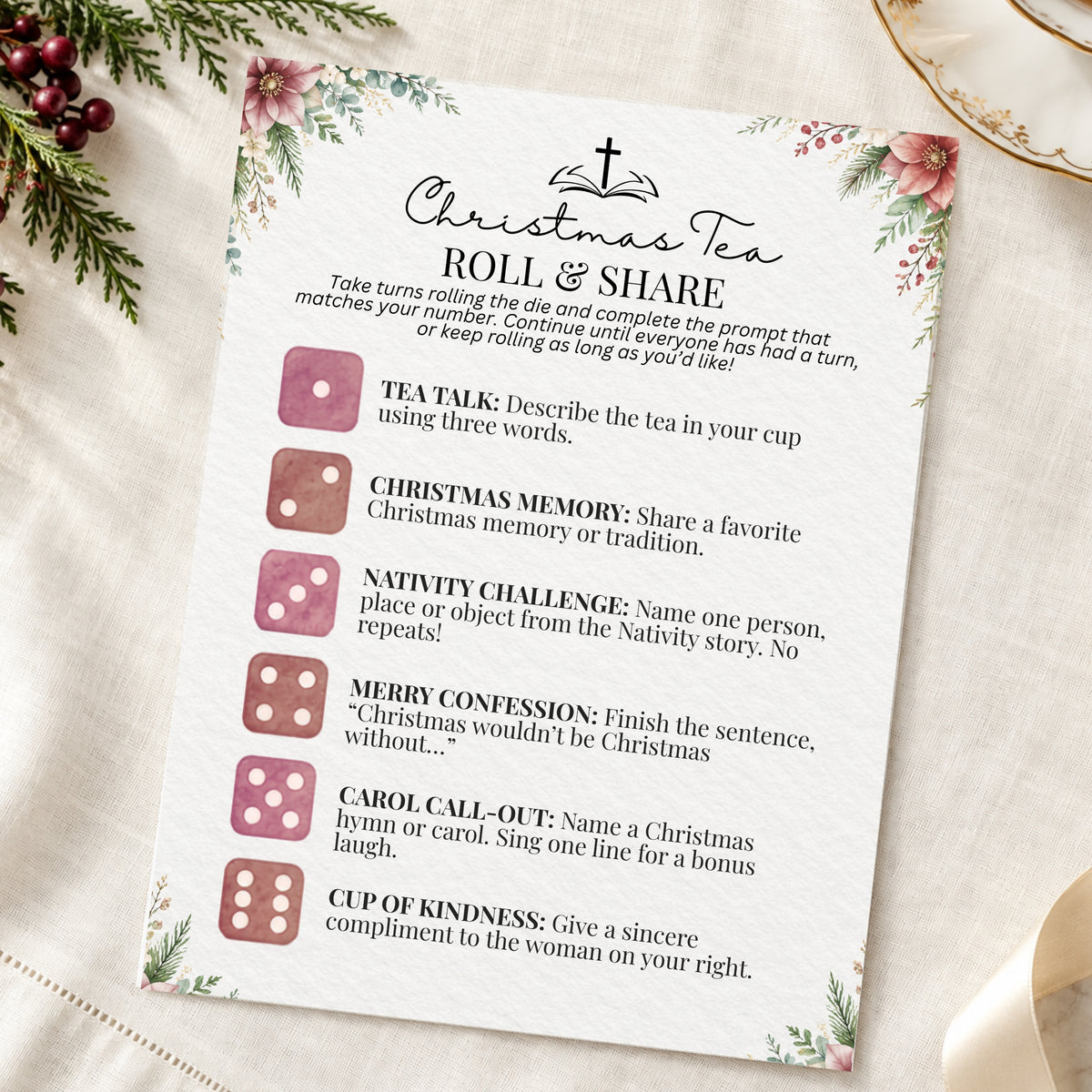 A printed Christmas tea game card titled "Roll & Share" with floral corner illustrations and pink dice icons, photographed on a cream fabric surface with holiday greenery and a gold plate edge visible around it.